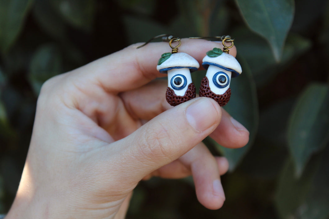 Two White and Blue Mushroom Earrings with Evil Eye, Leaves, and Textured Dirt.